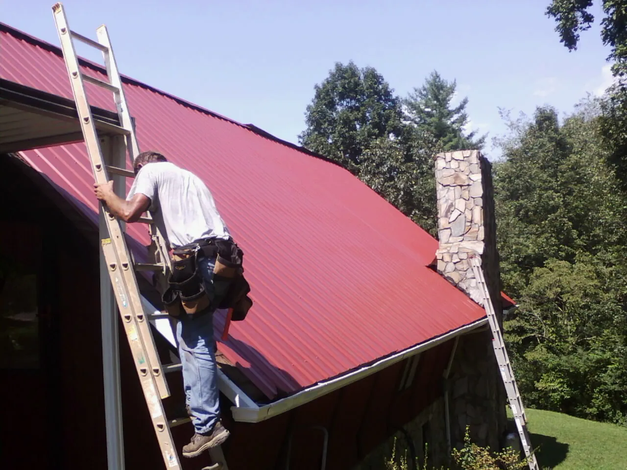 Roofer on a ladder beside a red metal roof and stone chimney — Soffit & Fascia Repair by Roofing Brothers in Luke Air Force Base
